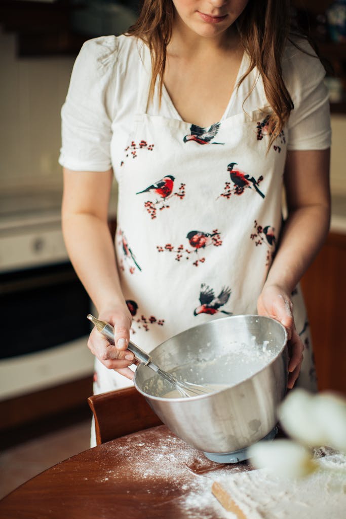 A woman in a kitchen mixing dough in a bowl while wearing a bird-patterned apron.